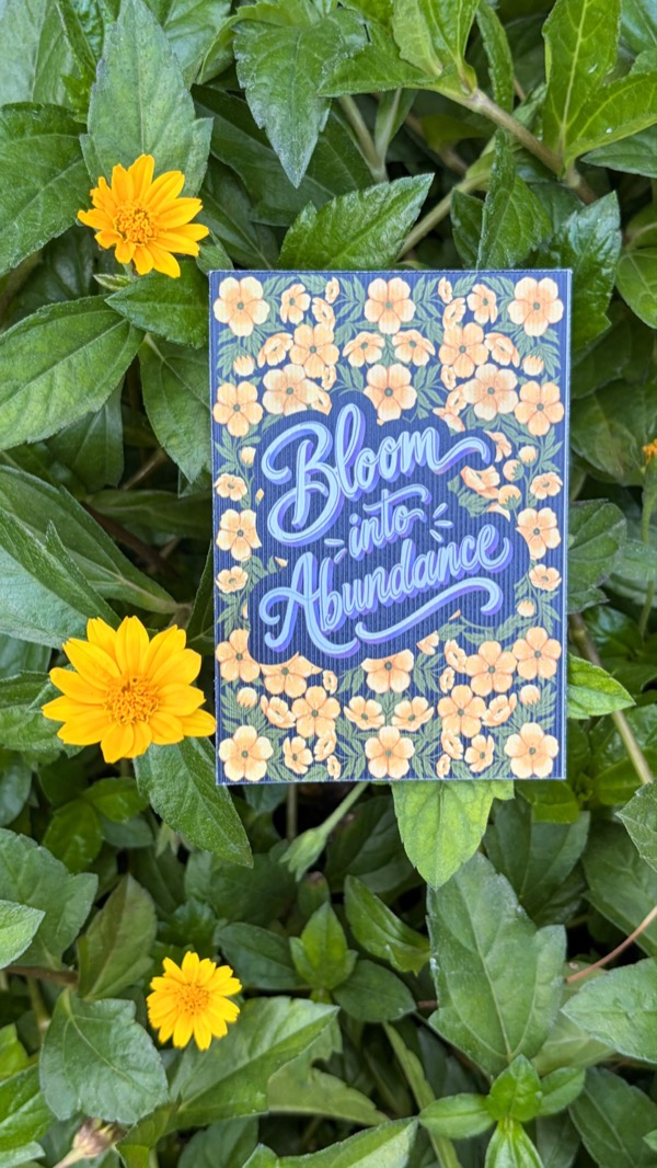 A blue lettered card reading “Bloom into Abundance” in a border of cream flowers, on green leaves with yellow flowers