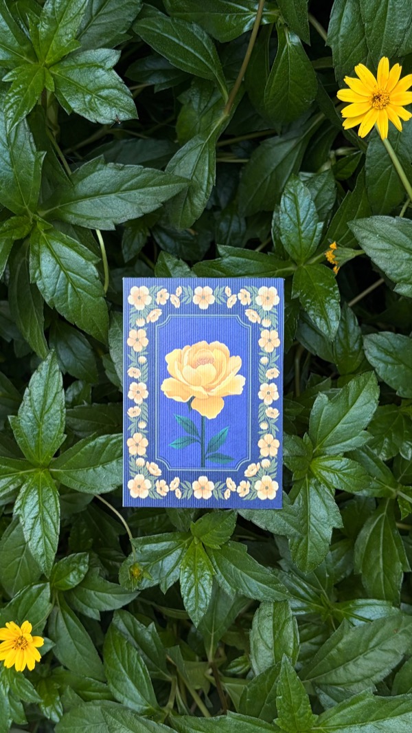 A blue mini print of a yellow peony inside a border of small cream flowers, resting on green leaves