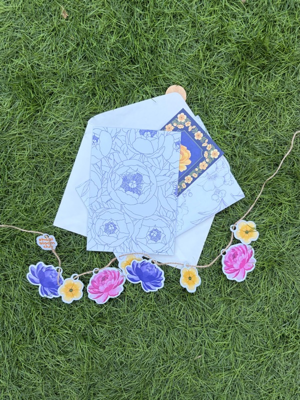 A peony colouring page on white envelopes on grass, with a garland of paper peonies strung across