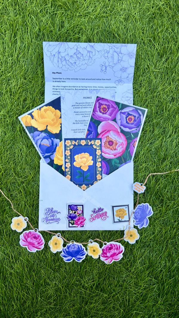 An open white envelope on grass with the letter, peony art prints and a blue mini print fanned out of it, floral stickers on its front, and a garland of paper peonies below
