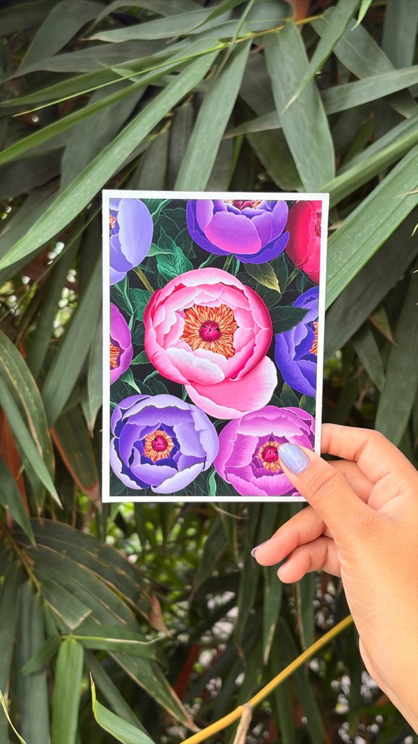 A hand holding a pink and purple peony art print against bamboo leaves